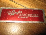 Reklameskilt i blikk, P.I.LANGLOS FABRIKKER, STRANDA. Ca 5.5 cm langt.
